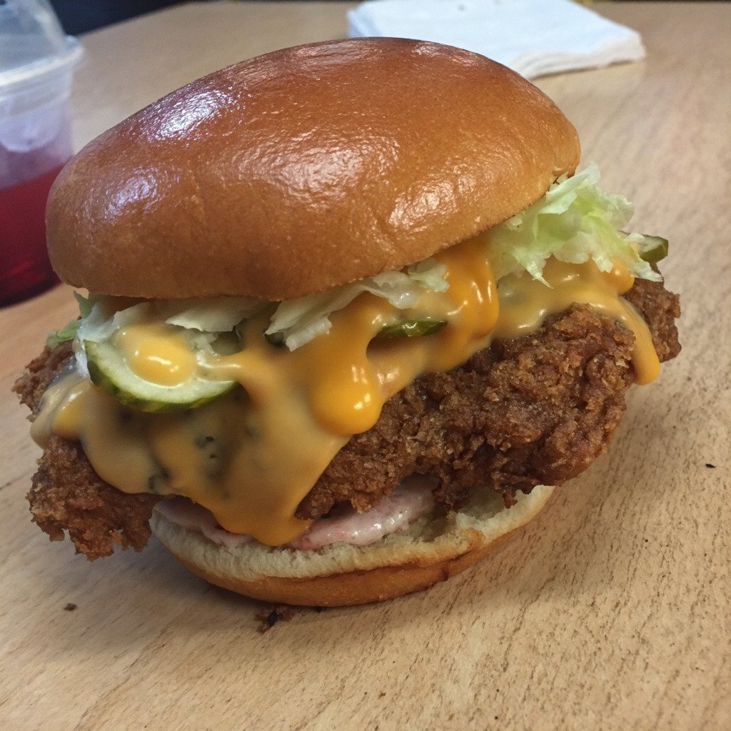 A phone photo of a fried chicken sandwich with cheese sauce, pickles and lettuce on a wooden table