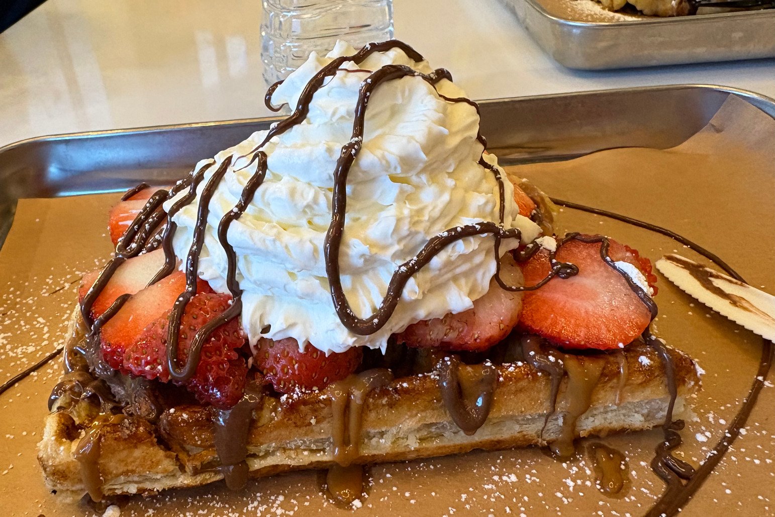 Photo: a waffle with strawberries, whipped cream and chocolate sauce on a metal tray